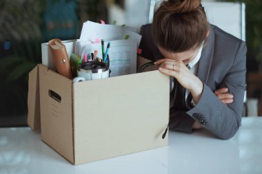 Yeni bir iş. Modern yeşil ofiste, gri takım elbiseli, karton kutuda kişisel eşyaları olan mutsuz bir modern kadın işçi..