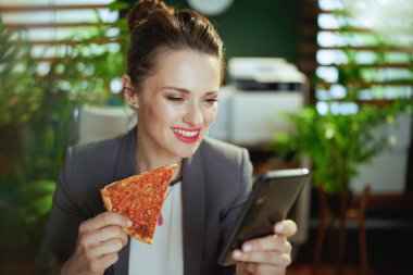 Sürdürülebilir iş yeri. Modern, 40 yaşında, modern yeşil ofisteki gri takım elbiseli, akıllı telefon uygulamaları kullanarak pizza yiyen küçük işletme sahibi bir kadın..