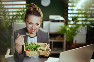 Sürdürülebilir iş yeri. Modern yeşil ofisteki gri takım elbiseli, dizüstü bilgisayarlı, salata yiyen, gülümseyen, küçük işletme sahibi kadın..