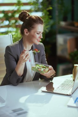 Sürdürülebilir iş yeri. Modern yeşil ofiste gri takım elbiseli, dizüstü bilgisayarı salata yiyen, gülümseyen modern kadın işçi..