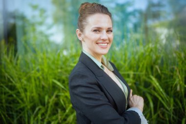 Orta yaşlı, gülümseyen bir iş kadınının portresi ofis binasının yanında. Siyah ceketli, kollarını kavuşturmuş..