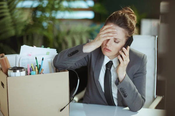 New job. sad modern female worker in modern green office in grey business suit with personal belongings in cardboard box talking on a smartphone.