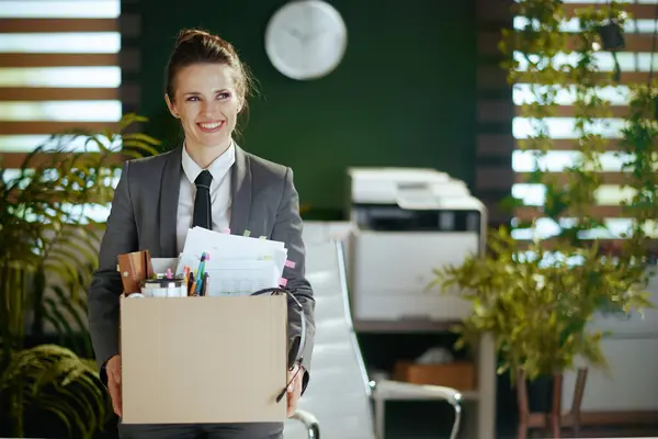 New job. smiling modern female employee in modern green office in grey business suit with personal belongings in cardboard box.