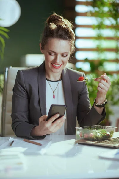 Sustainable workplace. smiling modern middle aged small business owner woman in a grey business suit in modern green office with laptop and smartphone eating salad.
