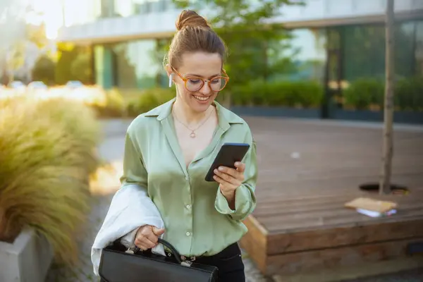 商务中心附近快乐的中年妇女 身穿绿色衬衫 戴着眼镜 头戴手提箱 手持无线耳机 用智能手机 — 图库照片