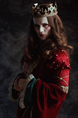 Kırmızı elbiseli, beyaz makyajlı ve tacı koyu gri arka planda korkunç bir ortaçağ kraliçesi..