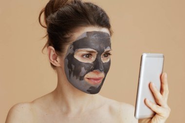 Bej renkte izole edilmiş akıllı telefon kullanan yüz maskeli genç bir kadın..