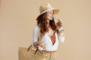 Plaj tatili Beyaz bluzlu mutlu orta yaşlı ev hanımı ve bej arka planda bir şişe su, saman torbası ve hasır şapkalı bir şort..