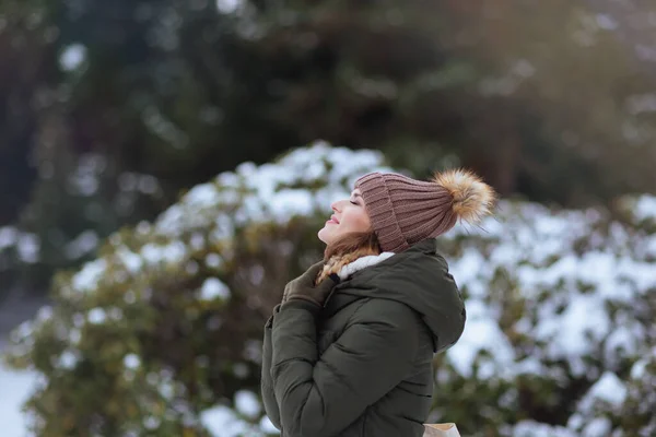 smiling modern woman in green coat and brown hat outdoors in the city park in winter with beanie hat near snowy branches.