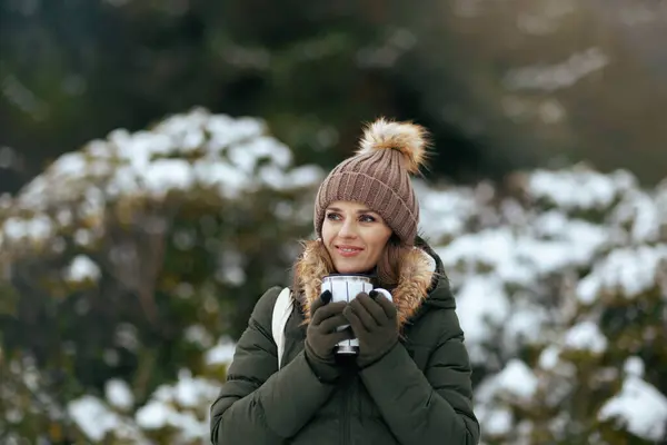 happy modern woman in green coat and brown hat outdoors in the city park in winter with mittens, cup of coffee and beanie hat.