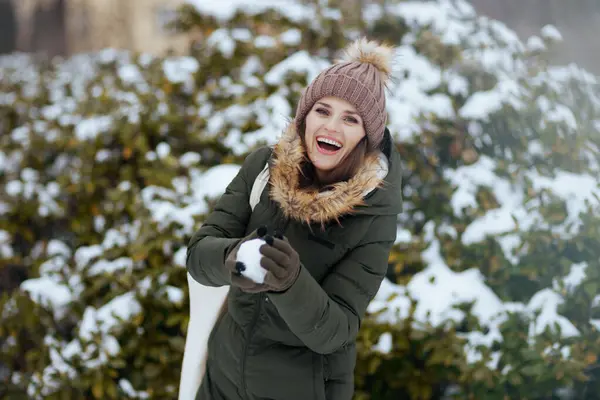 smiling modern woman in green coat and brown hat outdoors in the city park in winter with mittens and beanie hat throwing snowballs near snowy branches.