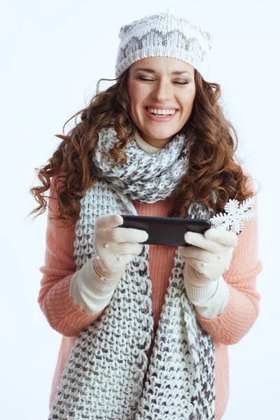 Hello winter. cheerful modern woman in sweater, mittens, hat and scarf against white background with snowflake sending text message using smartphone.