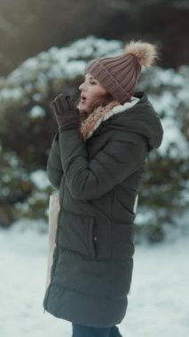 Kırk yıllık modern kadın yeşil ceketli ve kahverengi şapkalı kışın şehir parkında eldivenli ve bereli soğuk elleri nefesle ısıtan bir kadın..