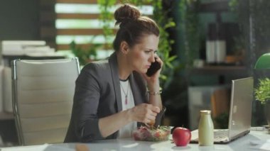 Sürdürülebilir iş yeri. Modern yeşil ofisteki gri takım elbiseli, modern orta yaşlı kadın işçi. Laptop yiyor ve akıllı telefondan konuşuyor..
