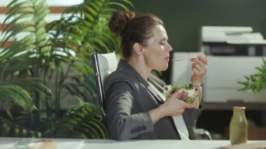 Sürdürülebilir iş yeri. Gri takım elbiseli, yeşil ofisteki kadın çalışanın arkasından salata yerken görülmüş..