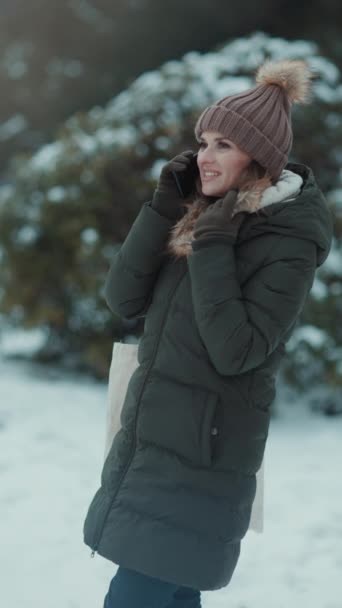 スマートフォンで話している雪の枝の近くのミトンとビーニー帽子と冬の市公園の緑のコートと茶色の帽子で現代40歳の女性を微笑みます — ストック動画