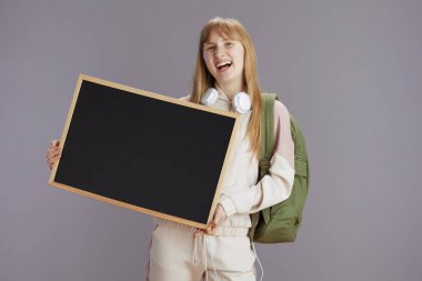 Sırt çantalı ve kulaklıklı bej eşofmanlı gülümseyen modern genç kız gri üzerinde boş tahtayı gösteriyor..