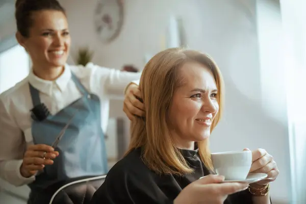 40 years old hair salon employee in modern beauty salon cutting hair with scissors and relaxed client with cup of tea.