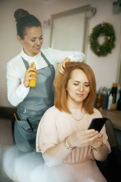 middle aged woman hairdresser in modern beauty salon with hair oil bottle and client with smartphone.