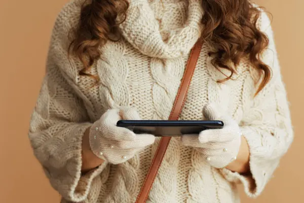 Hello winter. Closeup on woman in beige sweater, mittens and hat on beige background using smartphone applications.