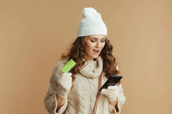 Hello winter. excited elegant woman in beige sweater, mittens and hat against beige background with smartphone and credit card buying on internet.