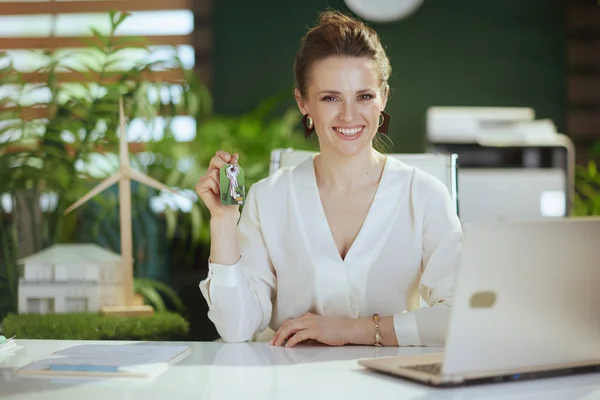 Sustainable real estate business. happy modern 40 years old woman realtor in modern green office in white blouse with laptop and keys.