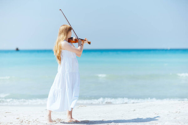 Full length portrait of modern young woman in white dress with violin enjoying playing on the beach.