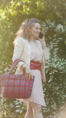 Mutlu, orta yaşlı, pembe elbiseli, beyaz ceketli bir kadın akıllı telefondan konuşuyor..