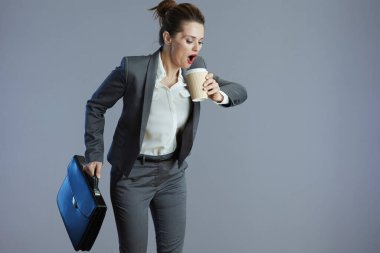 Gri takım elbiseli, kahve fincanı ve gri arka planda izole bir şekilde duran evrak çantasıyla şık bir kadın işçi..