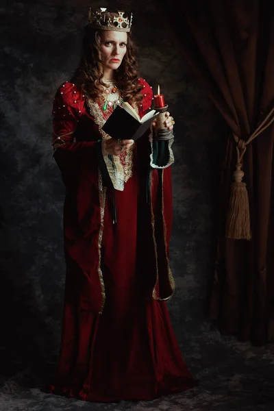 Full length portrait of medieval queen in red dress with book, candle and crown on dark gray background.