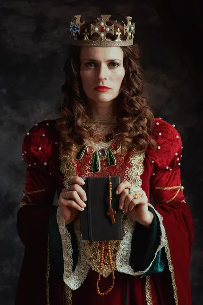 medieval queen in red dress with book, rosary and crown on dark gray background.