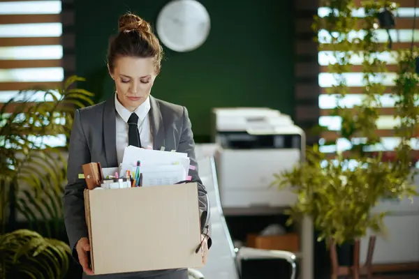 New job. sad modern woman worker in modern green office in grey business suit with personal belongings in cardboard box.