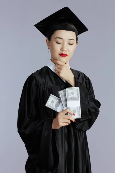 smiling modern female asian graduate student with money isolated on gray background.