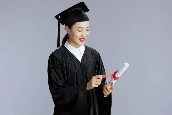 happy young female asian graduate student with diploma isolated on gray background.