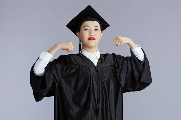 proud modern graduate student asian woman in graduation gown with cap showing biceps against grey background.