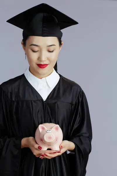 modern female asian graduate student with piggy bank against gray background.