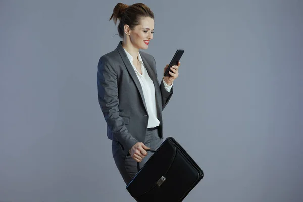 happy stylish small business owner woman in gray suit with smartphone and briefcase isolated on gray background.