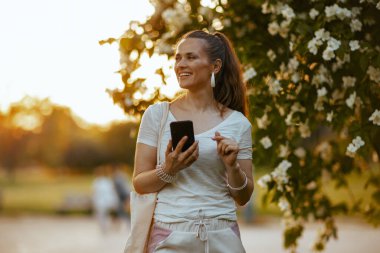 Yaz zamanı. Beyaz tişörtlü mutlu orta yaşlı kadın çiçek açan ağacın yanında akıllı telefon uygulaması kullanıyor..