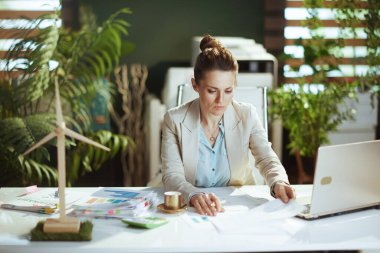 Modern yeşil ofisteki hafif takım elbiseli modern kadın işçi. Belgeler ve dizüstü bilgisayarla..