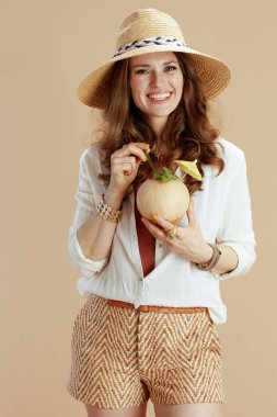 Plaj tatili Beyaz bluz ve bej arka planda şort giyen, hindistan cevizi kokteyli ve hasır şapkalı mutlu stil sahibi bir kadın..