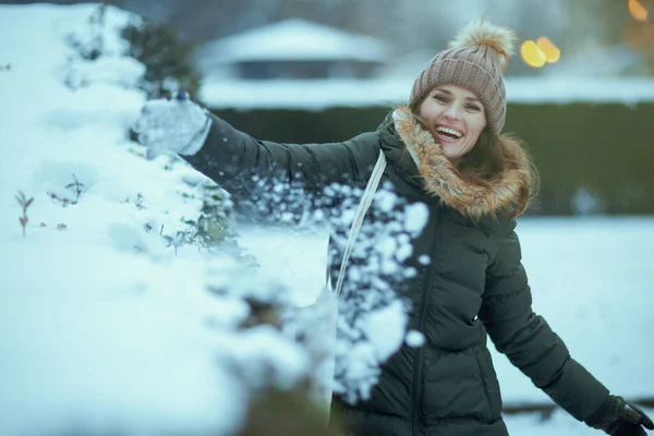 smiling modern woman in green coat and brown hat outdoors in the city park in winter with mittens and beanie hat playing with snow.