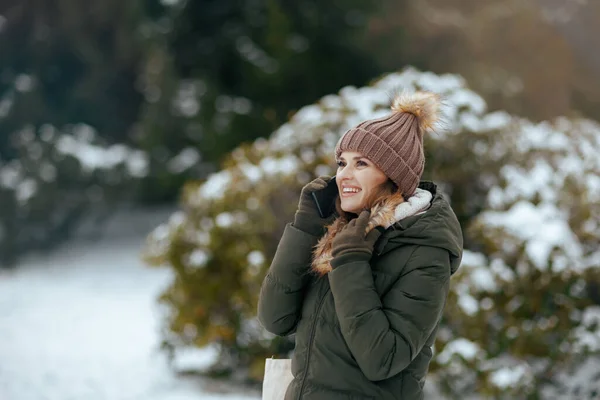 happy modern woman in green coat and brown hat outdoors in the city park in winter with mittens and beanie hat near snowy branches talking on a smartphone.