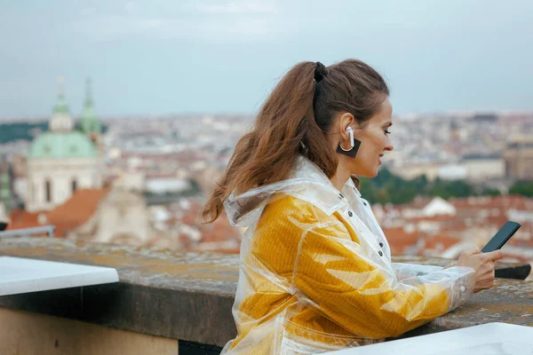 pensive young solo traveller woman in yellow blouse and raincoat in Prague Czech Republic with smartphone and headphones against city panorama.