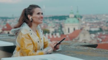 Prag 'da sarı bluz ve yağmurluk giyen, kulaklıklı, şehir panoramasına karşı akıllı telefon kullanan rahat genç bir kadın..
