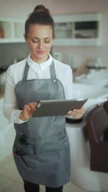 Modern güzellik salonundaki mutlu kadın kuaför. Üniformalı. Salon yıkama ünitesinin yanında tablet bilgisayar kullanıyor..