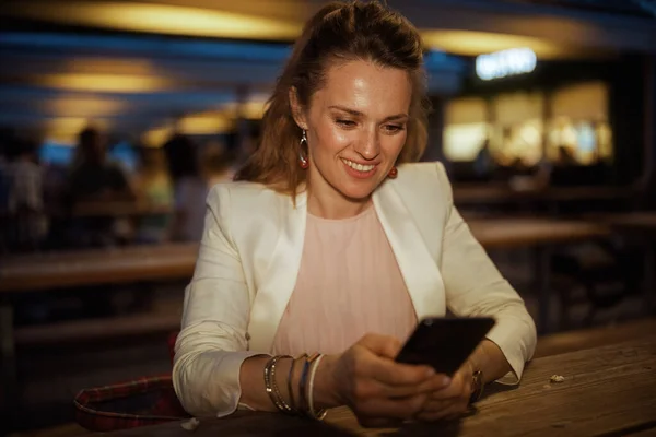 happy trendy woman in pink dress and white jacket in the city using smartphone app at the street cafe.