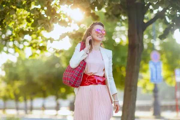 城市里穿着粉色衣服和白色夹克 头戴红包 戴着太阳镜 头戴智能手机的时尚女性 — 图库照片