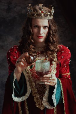 Kırmızı elbiseli sinsi ortaçağ kraliçesi koyu gri arka plandaki kadehe zehir döktü..