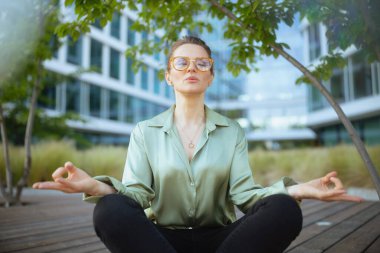 Rahatlamış modern kadın işçi ofis binasının yanında yeşil bluz ve gözlüklerle meditasyon yapıyor..