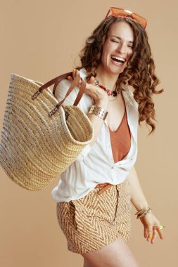Plaj tatili Beyaz bluz ve şortla gülümseyen, saman torbası ve güneş gözlüklü, bej arkaplan.